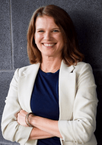 Headshot of Kristen Evans smiling, wearing navy top and neutral blazer with arms crossed against a gray brick wall